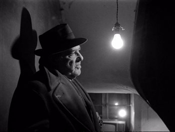 Movie still from “Alphaville” (1965), directed by Jean-Luc Godard – A man in a hat standing in front of a light; Medium shot, Low angle