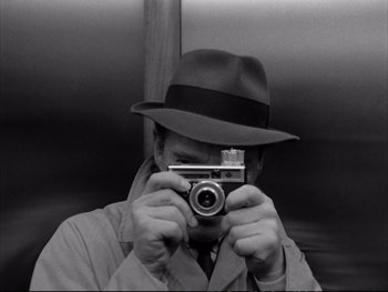Movie still from “Alphaville” (1965), directed by Jean-Luc Godard – A black and white photo of a man taking a picture; Close Up shot, High angle