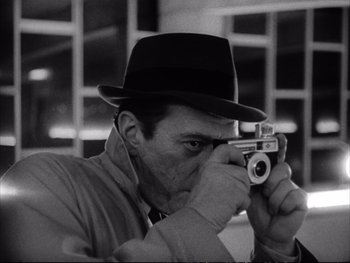 Movie still from “Alphaville” (1965), directed by Jean-Luc Godard – A black and white photo of a man taking a picture with a camera; Close Up shot, Low angle