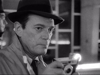 Movie still from “Alphaville” (1965), directed by Jean-Luc Godard – A black and white photo of a man holding a gun; Close Up shot, Low angle