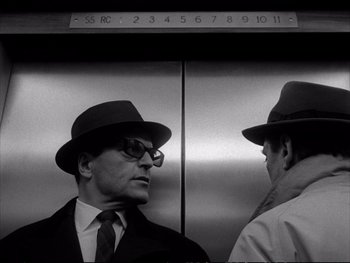 Movie still from “Alphaville” (1965), directed by Jean-Luc Godard – A man in a suit and a hat standing next to another man in an elevator; Close Up shot, Low angle