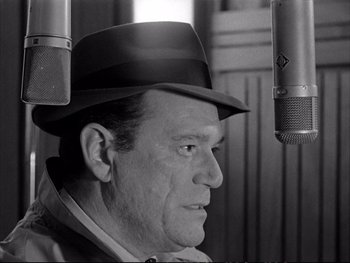 Movie still from “Alphaville” (1965), directed by Jean-Luc Godard – An older man wearing a hat in front of a microphone; Close Up shot, Low angle