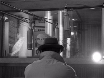 Movie still from “Alphaville” (1965), directed by Jean-Luc Godard – A man wearing a hat looking at a microphone; Medium shot, Over the shoulder angle