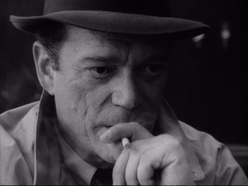 Movie still from “Alphaville” (1965), directed by Jean-Luc Godard – An older man wearing a hat and smoking a cigarette; Close Up shot, Low angle