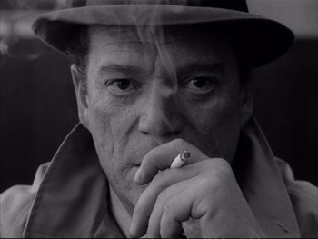 Movie still from “Alphaville” (1965), directed by Jean-Luc Godard – A black and white photo of a man smoking a cigarette; Close Up shot, Low angle