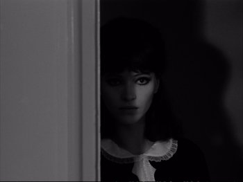 Movie still from “Alphaville” (1965), directed by Jean-Luc Godard – A black - and - white photo of a woman in front of a door; Close Up shot, Over the shoulder angle
