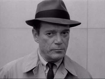 Movie still from “Alphaville” (1965), directed by Jean-Luc Godard – An older man wearing a hat and a trench coat; Close Up shot, High angle
