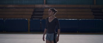 Movie still from “Alps” (2011), directed by Yorgos Lanthimos – A girl in a black shirt is standing in a gym; Medium shot, Over the shoulder angle