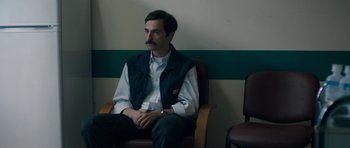 Movie still from “Alps” (2011), directed by Yorgos Lanthimos – A man sitting in a chair with his hands crossed; Medium shot, Over the shoulder angle