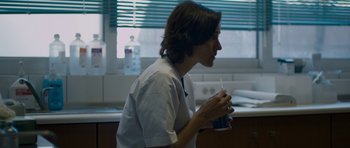 Movie still from “Alps” (2011), directed by Yorgos Lanthimos – A person holding a cup in a room; Medium shot, Over the shoulder angle