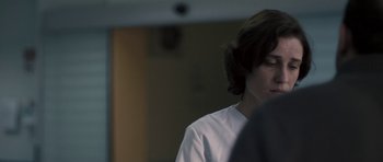 Movie still from “Alps” (2011), directed by Yorgos Lanthimos – A person with long hair is standing in a room; Close Up shot, Over the shoulder angle