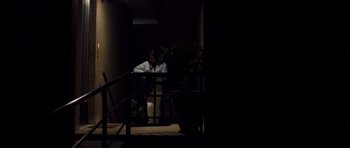 Movie still from “Alps” (2011), directed by Yorgos Lanthimos – A person sitting in a dark room next to a railing and potted plant; Wide shot, Low angle
