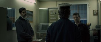 Movie still from “Alps” (2011), directed by Yorgos Lanthimos – A man wearing a hat in a room with other people; Medium shot, Over the shoulder angle