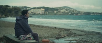 Movie still from “Alps” (2011), directed by Yorgos Lanthimos – A person sitting on a bench on the beach; Medium shot, Low angle