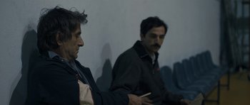 Movie still from “Alps” (2011), directed by Yorgos Lanthimos – Two men sitting next to each other in front of a white wall; Medium shot, Over the shoulder angle