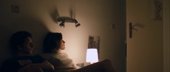 Movie still from “Alps” (2011), directed by Yorgos Lanthimos – A woman sitting on a bed with a lamp in a room; Medium shot, High angle