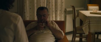 Movie still from “Alps” (2011), directed by Yorgos Lanthimos – An older man sitting in a chair drinking from a glass; Medium shot, High angle