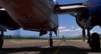 Movie still from “Always” (1989), directed by Steven Spielberg – A person riding a bike on a runway near an airplane; Extreme Wide shot, Low angle