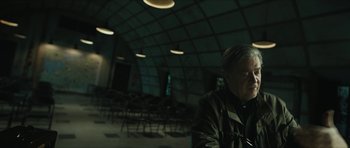 Movie still from “American Dharma” (2018), directed by Errol Morris – A man sitting in front of a group of chairs in a room; Medium shot, Over the shoulder angle