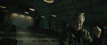 Movie still from “American Dharma” (2018), directed by Errol Morris – A man sitting in front of a group of chairs in a room; Medium shot, Over the shoulder angle