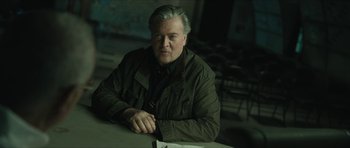 Movie still from “American Dharma” (2018), directed by Errol Morris – An older man sitting at a table in a dark room; Medium shot, Low angle