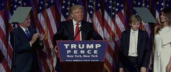 Movie still from “American Dharma” (2018), directed by Errol Morris – A man standing at a podium in front of american flags; Medium shot, Low angle