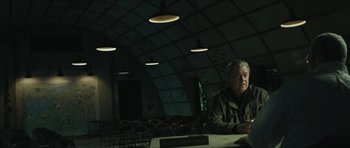 Movie still from “American Dharma” (2018), directed by Errol Morris – An older man sitting at a table in a dark room; Medium shot, Low angle