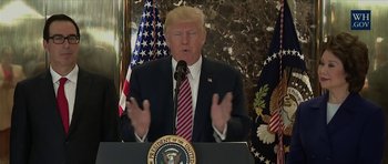 Movie still from “American Dharma” (2018), directed by Errol Morris – A man standing at a podium in front of two american flags; Medium shot, Over the shoulder angle