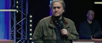 Movie still from “American Dharma” (2018), directed by Errol Morris – A man in a green jacket is holding a microphone; Close Up shot, Low angle