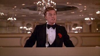 Movie still from “American Gigolo” (1980), directed by Paul Schrader – A man in a tuxedo standing at a podium in front of an audience; Medium shot, Low angle