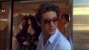 Movie still from “American Gigolo” (1980), directed by Paul Schrader – A man wearing sunglasses standing in front of a group of people; Close Up shot, Over the shoulder angle