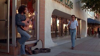 Movie still from “American Gigolo” (1980), directed by Paul Schrader – A man walking down a street while a man plays guitar; Wide shot, Over the shoulder angle
