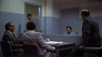 Movie still from “American Gigolo” (1980), directed by Paul Schrader – A man sitting at a table in front of two other men; Wide shot, Over the shoulder angle
