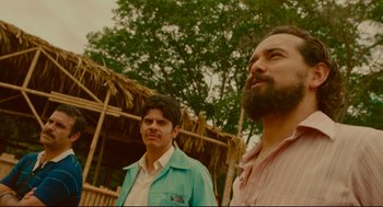 Movie still from “American Made” (2017), directed by Doug Liman – Two men standing next to each other in front of a hut; Medium shot, Low angle
