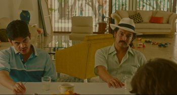 Movie still from “American Made” (2017), directed by Doug Liman – A man in a hat sitting at a table with two other people; Medium shot, Over the shoulder angle