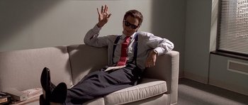 Movie still from “American Psycho” (2000), directed by Mary Harron – A man sitting on top of a couch waving; Medium shot, Low angle
