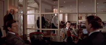 Movie still from “American Psycho” (2000), directed by Mary Harron – A man sitting at a table in front of a glass wall; Wide shot, Over the shoulder angle