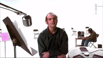 Movie still from “American Splendor” (2003), directed by Shari Springer Berman – A man sitting in front of a microphone in front of a camera; Medium shot, Over the shoulder angle