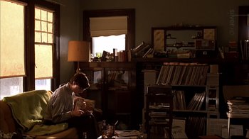 Movie still from “American Splendor” (2003), directed by Shari Springer Berman – A man sitting in front of a bookshelf reading a book; Wide shot, High angle