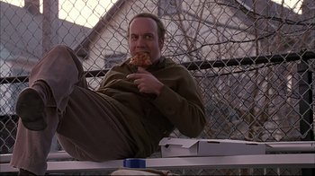 Movie still from “American Splendor” (2003), directed by Shari Springer Berman – A man eating a piece of pizza while sitting on top of a bench; Medium shot, Low angle