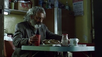 Movie still from “Amores Perros” (2000), directed by Alejandro G. Iñárritu – An older man sitting at a table with food; Medium shot, Low angle