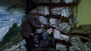 Movie still from “Amores Perros” (2000), directed by Alejandro G. Iñárritu – A man sitting in front of a pile of papers while another man combs his hair; Medium shot, High angle