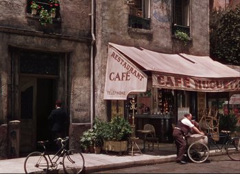 Movie still from “An American in Paris” (1951), directed by Vincente Minnelli – A man riding a bike down a street next to a cafe; Extreme Wide shot, High angle