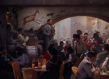Movie still from “An American in Paris” (1951), directed by Vincente Minnelli – A group of people sitting around a room with a man playing a saxophone; Wide shot, High angle