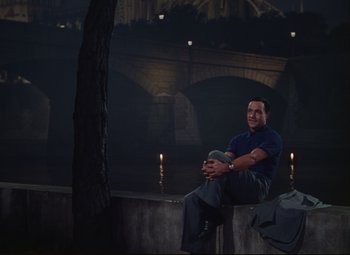 Movie still from “An American in Paris” (1951), directed by Vincente Minnelli – A man sitting on a wall in front of a bridge; Wide shot, High angle