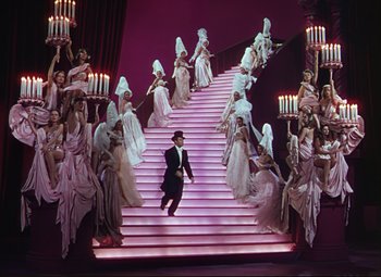 Movie still from “An American in Paris” (1951), directed by Vincente Minnelli – A man walking down a set of stairs surrounded by a group of women; Wide shot, High angle