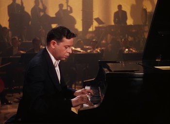 Movie still from “An American in Paris” (1951), directed by Vincente Minnelli – A man in a black suit playing a black piano in front of an orchestra; Medium shot, High angle