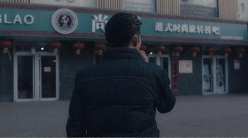 Movie still from “An Elephant Sitting Still” (2018), directed by Bo Hu – A man in a black jacket talking on a cell phone in front of a building; Medium shot, Over the shoulder angle