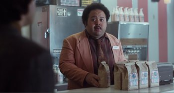 Movie still from “An Evening with Beverly Luff Linn” (2018), directed by Jim Hosking – A man standing at a counter with two bags of food; Medium shot, Over the shoulder angle