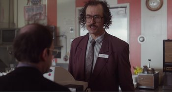 Movie still from “An Evening with Beverly Luff Linn” (2018), directed by Jim Hosking – A man with curly hair wearing a suit and tie talking to another man in front of him; Medium shot, Over the shoulder angle